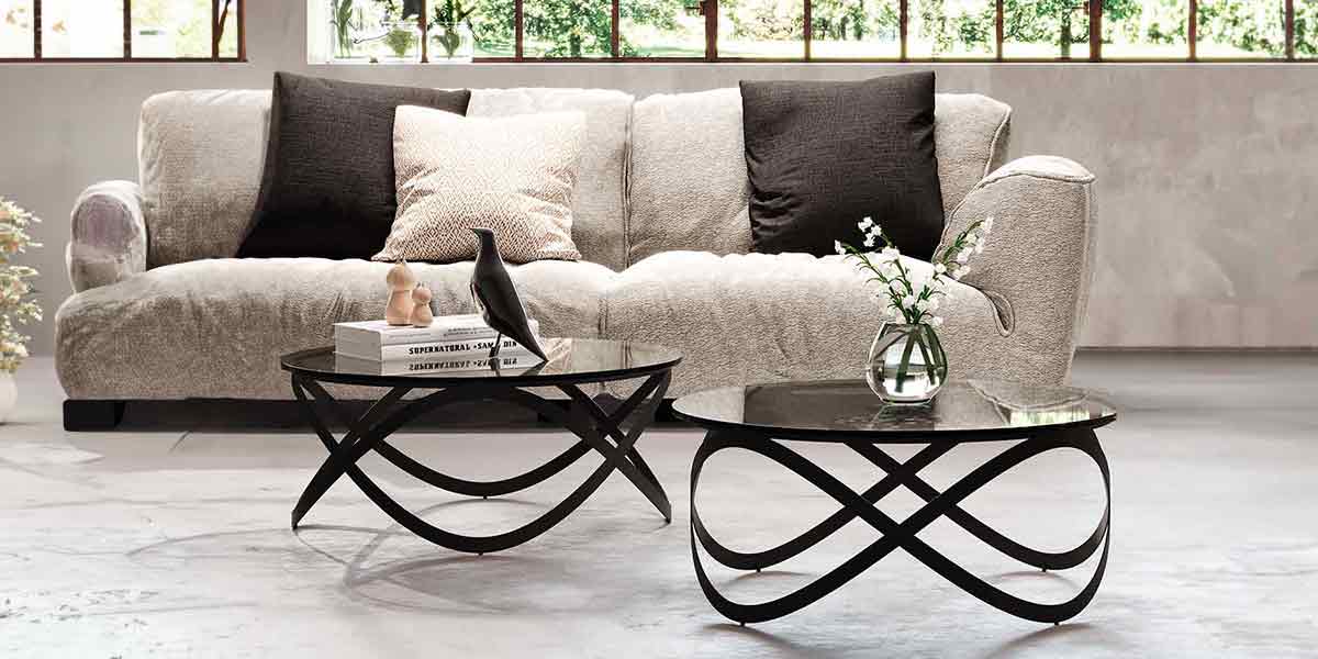 Kategorie Banner Couchtisch Candy K 900 mit Tischplatte Parsolglas grau und schwarzem Gestell Zwei Couchtische mit dunkler runder Glastischplatte stehen nebeneinander. Das geschwungene Gestell aus zwei Metallbändern sieht aus jeder Perspektive unterschiedlich aus.